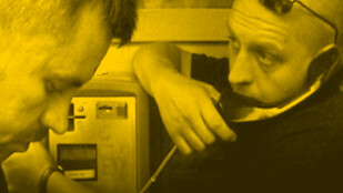 Czarno-żółte zdjęcie. Dwóch mężczyzn z profilu. Łysy mężczyzna po prawej trzyma w ręce słuchawkę i łokciem opiera się o budkę telefoniczną. Po prawej mężczyzna oprószony siwizną patrzy w dół.