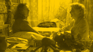 Czarno-żółte zdjęcie: Kobieta po czterdziestce i jej starsza mama siedzą naprzeciwko siebie. Na stole bałagan, leży na nim dużo przedmiotów. W tle sterta ubrań i zasłona. Zza zasłony prześwitują drzwi balkonowe i okno.