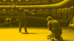 Czarno-żółte zdjęcie: Dwie kobiety na scenie w operze. Widok ze sceny na publiczność. Śpiewaczka operowa o krótkich, czerwonych włosach ma otwarte szeroko usta. Śpiewa z szeroko rozłożonymi rękoma. Przed nią kobieta klęczy na kolanach. Jest odwrócona tyłem do obiektywu.